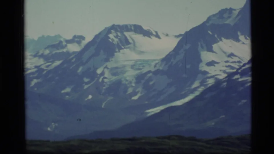 Wider 1977 Alaska 8mm view of snow-covered peaks, glacier-like ice, rocky slopes, and low green foreground terrain.