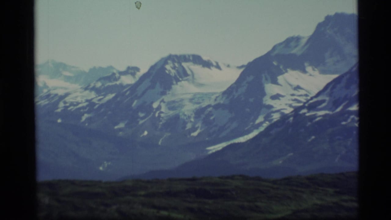 1977 Alaska 8mm frame showing blue-gray ridges and snow-covered peaks above a dark rolling foreground.