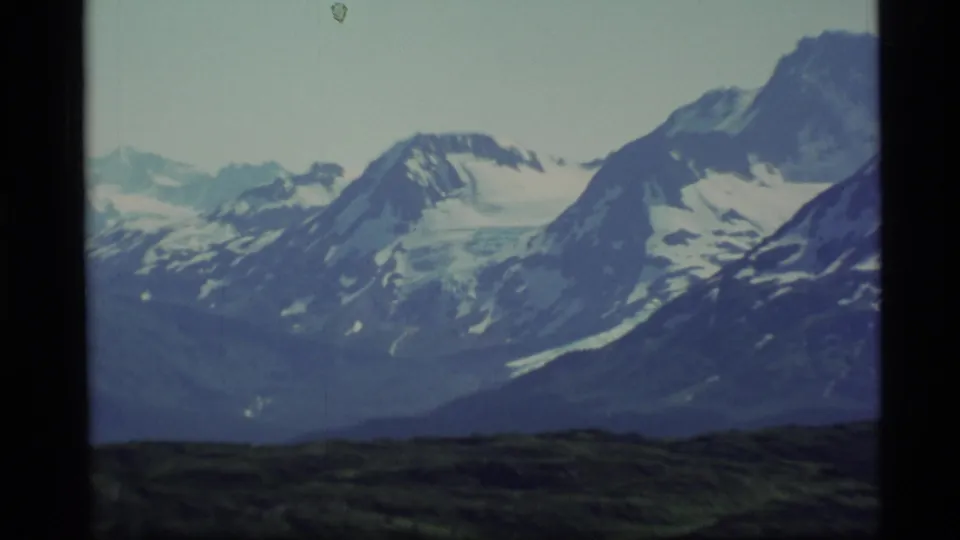 1977 Alaska 8mm frame showing blue-gray ridges and snow-covered peaks above a dark rolling foreground.