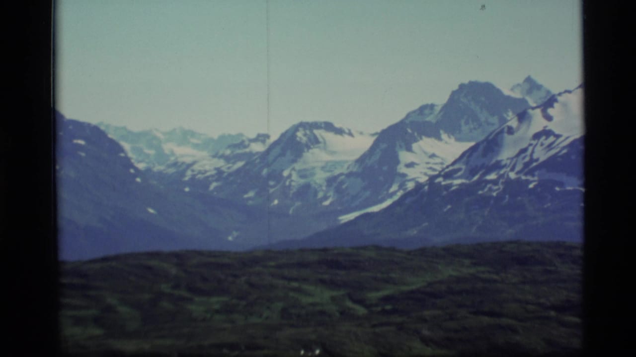 Wide mountain view with snow patches, pale sky, and dark foreground hills.