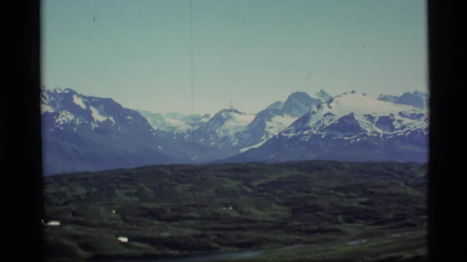 1977 Alaska archival frame of a snowy mountain range, dark tundra foreground, and pale sky with visible 8mm texture.