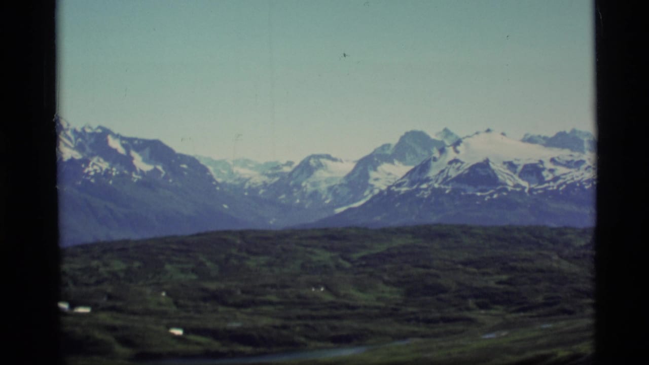 1977 Alaska 8mm frame showing distant snow-capped mountains, rolling dark foreground terrain, and black film borders.
