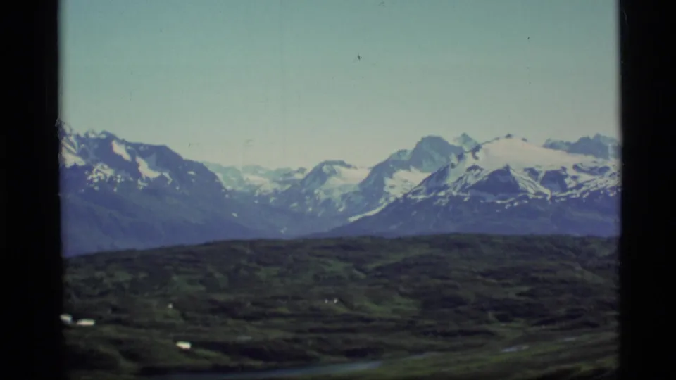 1977 Alaska 8mm frame showing distant snow-capped mountains, rolling dark foreground terrain, and black film borders.