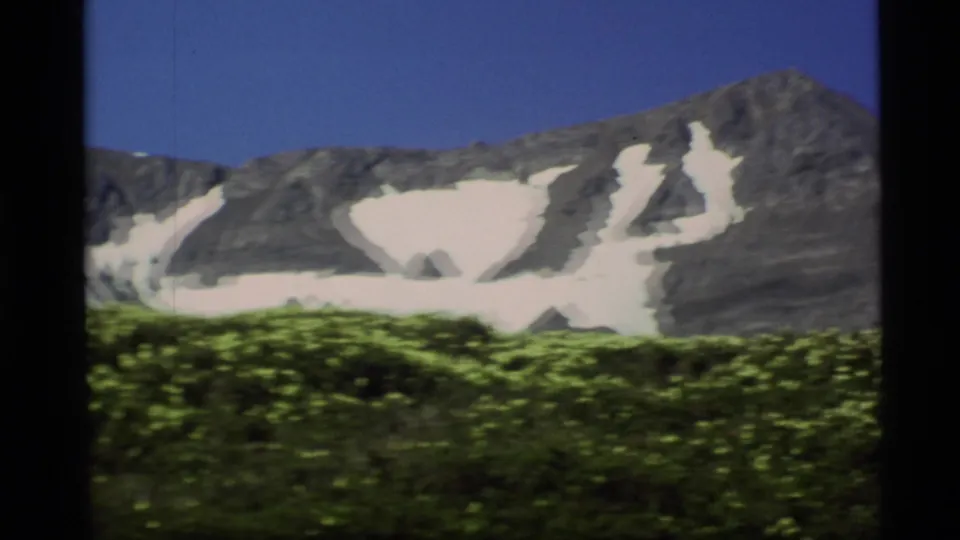 1977 Alaska 8mm frame with a blue sky, rocky mountain ridge, lingering snowfields, and yellow wildflowers in the lower frame.