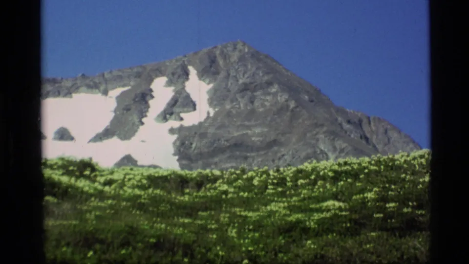 Alaska home-movie frame from 1977 with a rugged mountain ridge, bright snow patches, and dense green foreground vegetation.