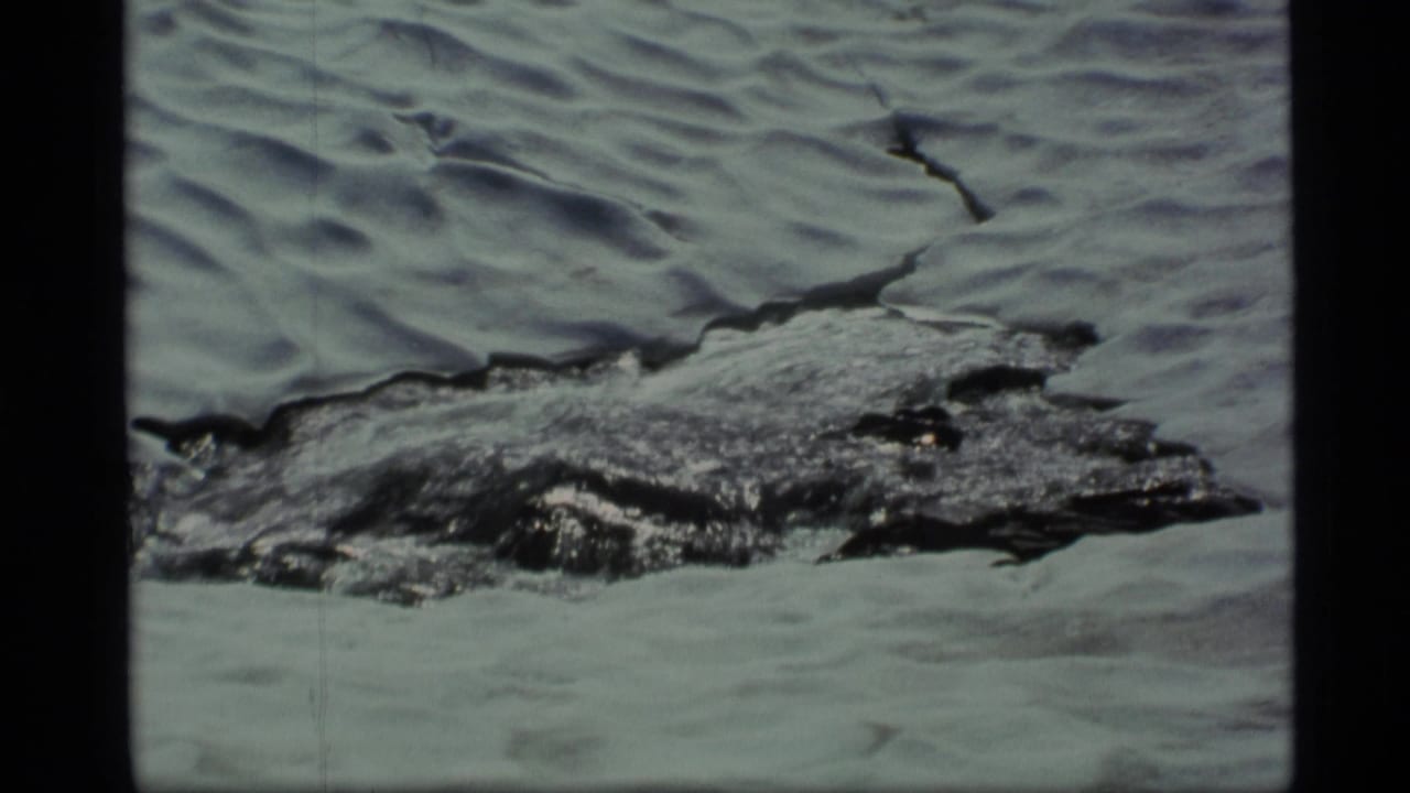1977 Alaska 8mm frame showing snow and ice around a dark channel of spring runoff in rocky mountain terrain.