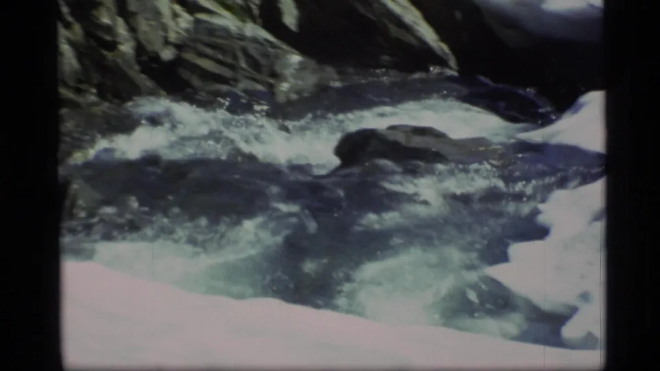 Vintage 8mm view of Alaskan snowmelt water moving through dark rocks beside a bright snowbank.
