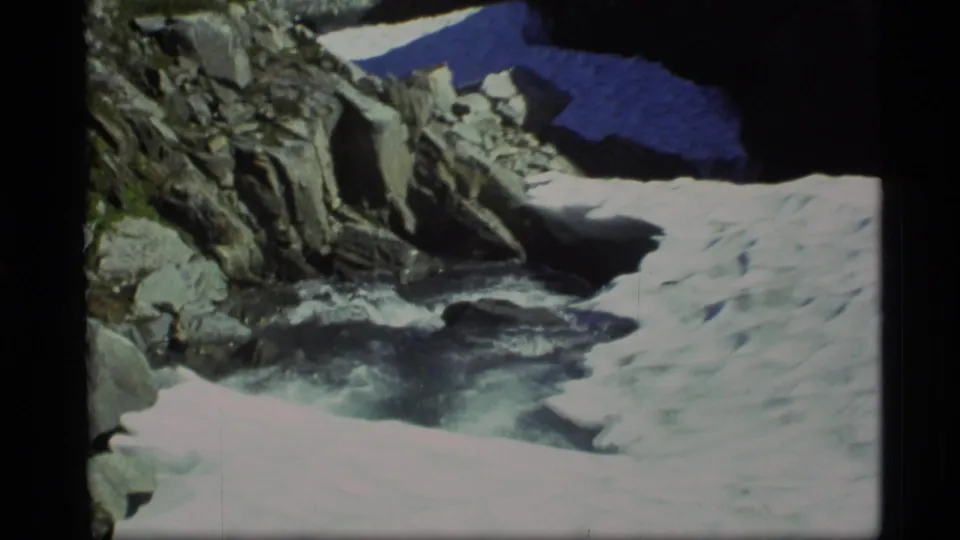 1977 Alaska 8mm frame of a rocky creek bordered by snowbanks and steep shaded stone.