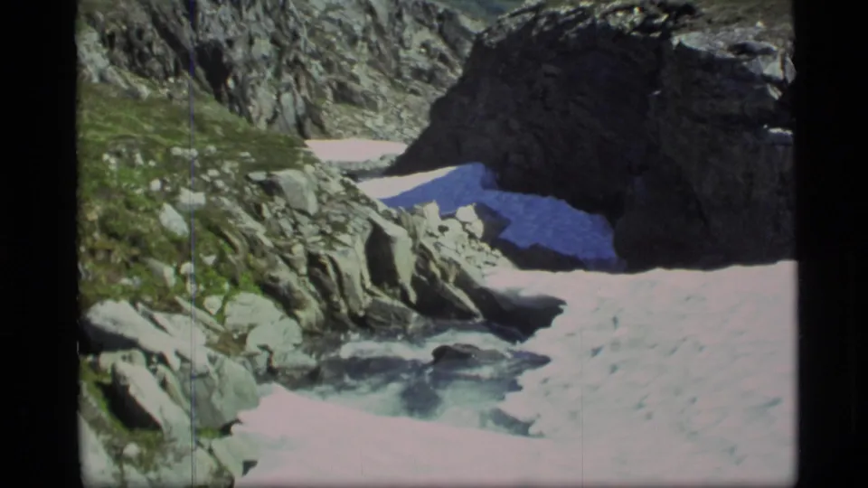 1977 Alaska home-movie frame showing a snow-edged mountain stream with rocks, green ground cover, and a shadowed cliff.