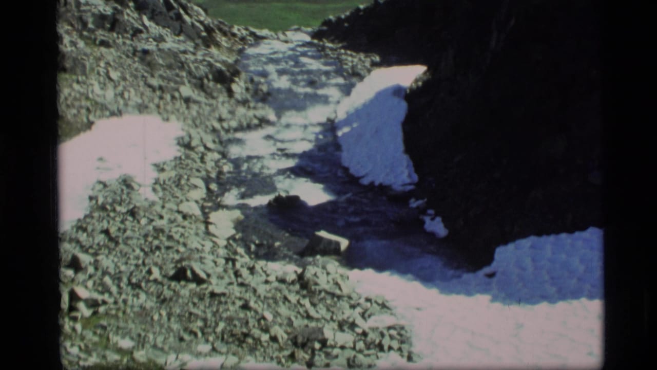 1977 Alaska 8mm frame showing a rocky ravine with melting snow patches and a stream running through the mountain terrain.
