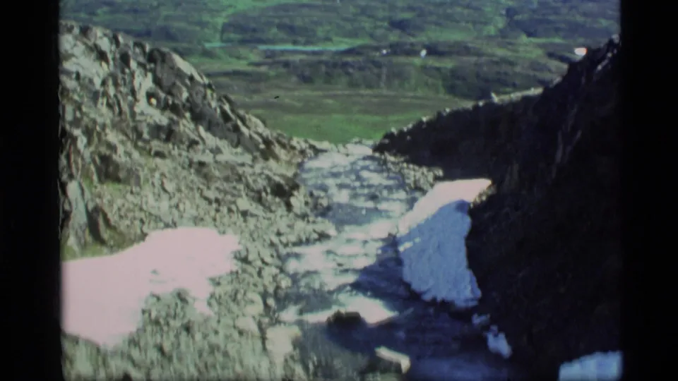 1977 Alaska 8mm frame looking down a rocky stream channel toward a green valley with distant snow-capped mountains.