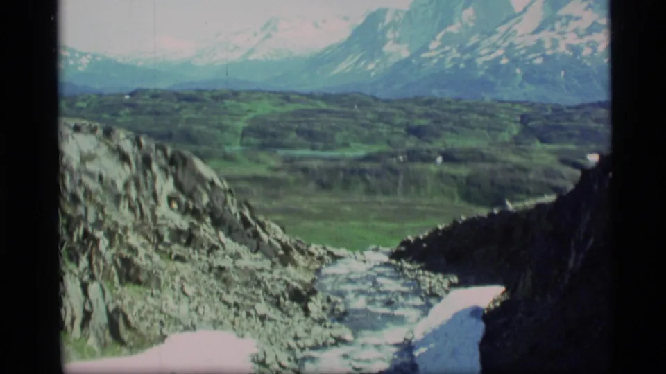 1977 Alaska 8mm frame showing a broader mountain view from a rocky ravine toward green lowlands and snow-capped peaks.