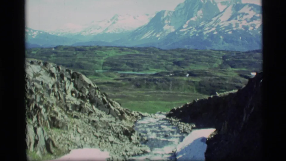 1977 Alaska 8mm frame with a wide view of a rocky snow-lined ravine leading toward a green valley and distant mountains.