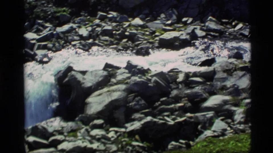 1977 Alaska 8mm frame of water rushing through a rocky streambed with bright foam and moss-covered stones.