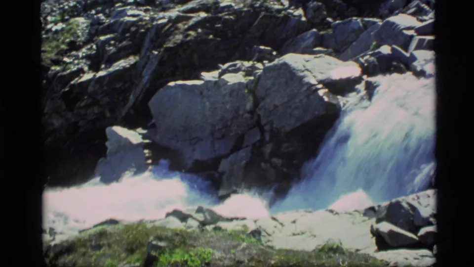 1977 Alaska home-movie frame with rushing water squeezed between angular rocks and sunlit foam.
