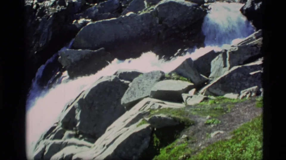 8mm Alaska frame of a rocky cascade with bright whitewater, boulders, and green moss along the lower edge.