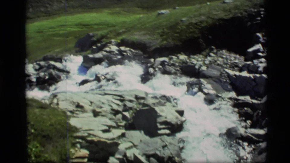 1977 Alaska 8mm frame showing a rocky stream running through green grass and uneven terrain.