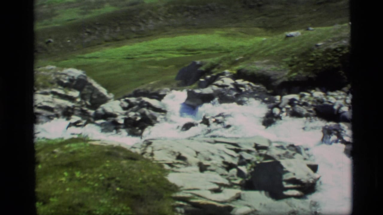 1977 Alaska 8mm frame showing a fast mountain stream crossing rocky terrain with grassy banks and clear sky.