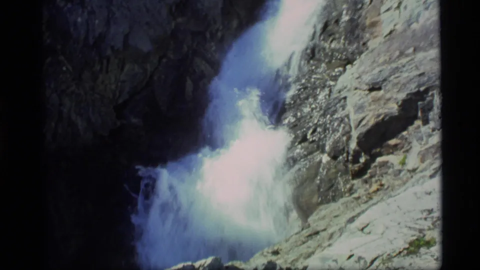 1977 Alaska 8mm frame showing churning whitewater at the base of steep gray rocks, with strong sun exposure.