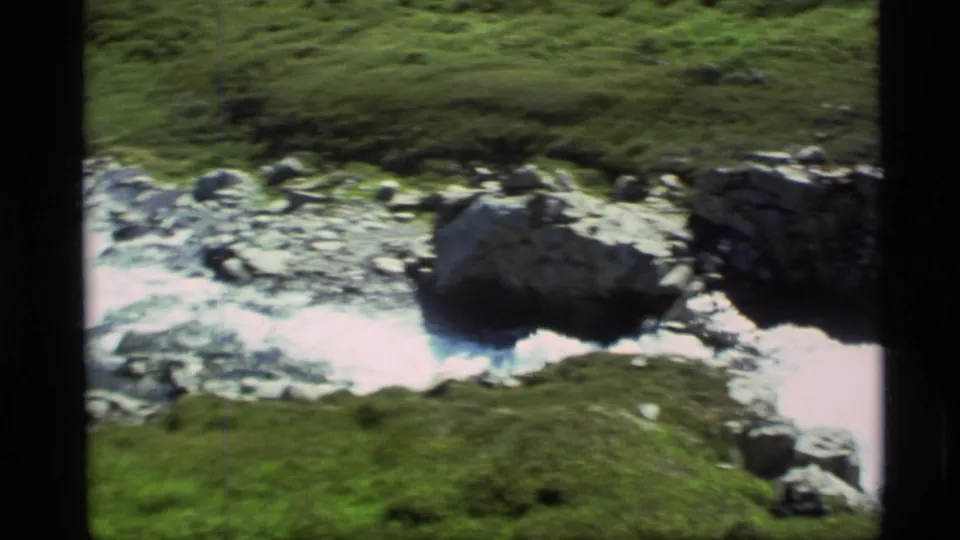 Vintage Alaska home-movie frame of water flowing around large rocks below a green slope.
