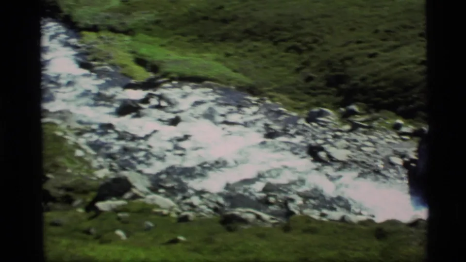 1977 8mm Alaska frame showing a fast stream spread across a rocky bed with green banks.