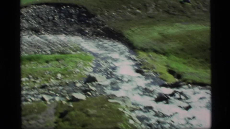 Archival 8mm frame from Alaska showing a narrow rocky channel with water and green terrain.