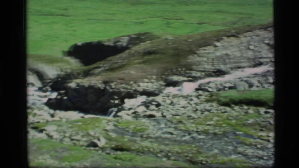 1977 Alaska home-movie frame of running water cutting through rocks and green valley ground.