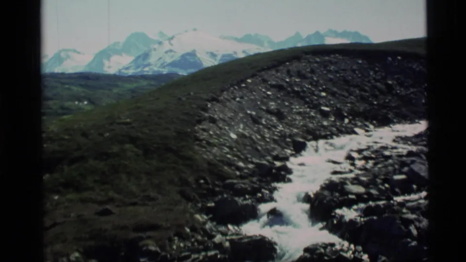 1977 Alaska 8mm frame with a rocky stream and distant snow-capped mountains in the background.