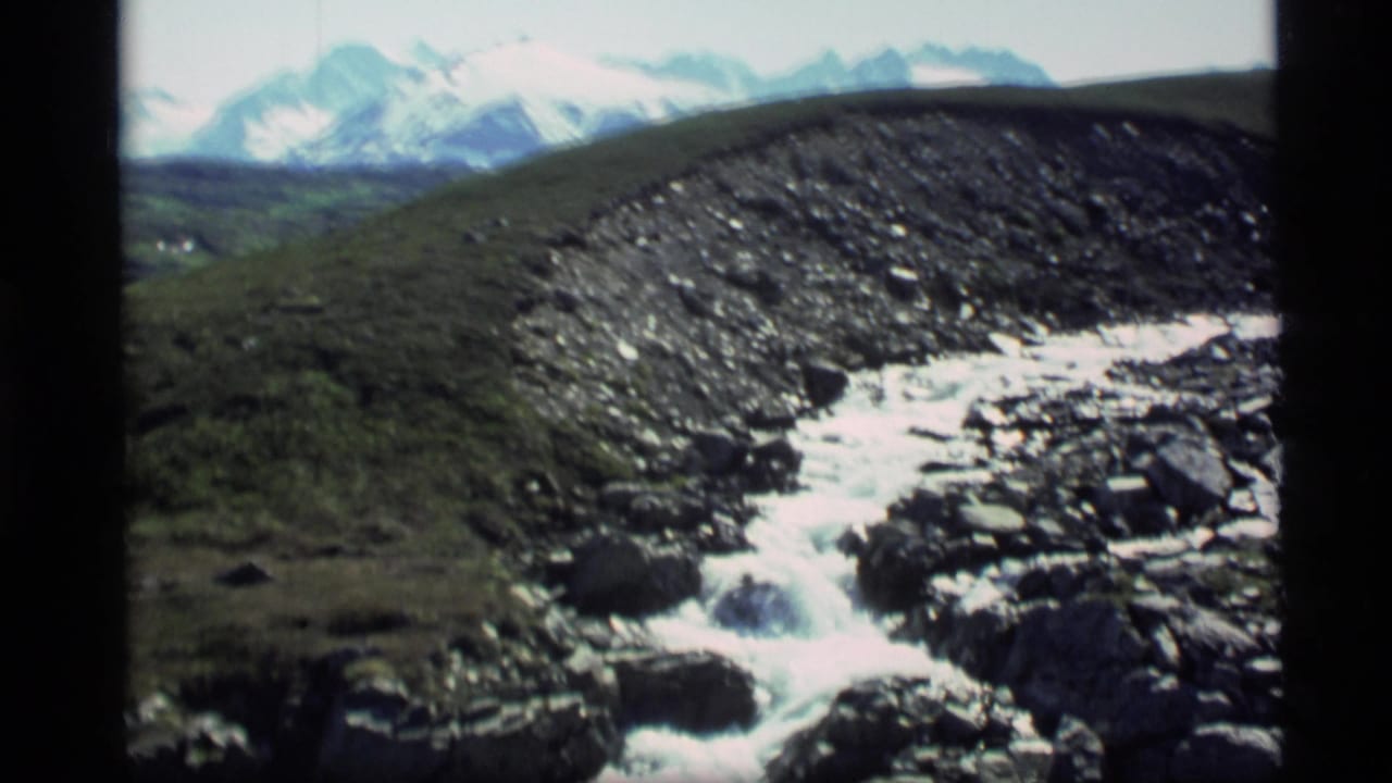 1977 Alaska 8mm frame of snowmelt water crossing a rocky slope, with distant snow-covered mountains in the background.