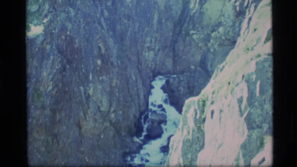 1977 Alaska home-movie frame looking down into a narrow rocky canyon with pale snowmelt water below.