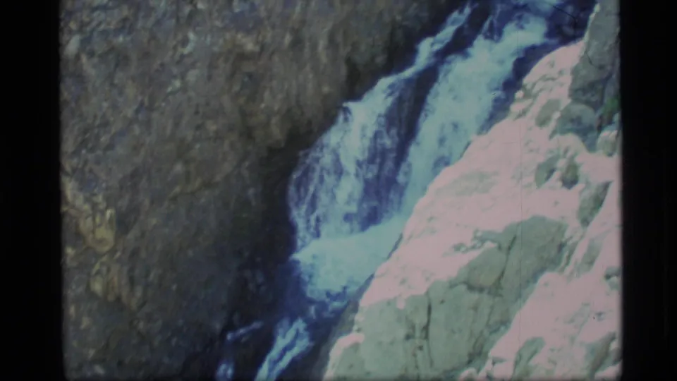 1977 Alaska home-movie frame with a waterfall descending beside pale rock ledges and shadowed canyon stone.