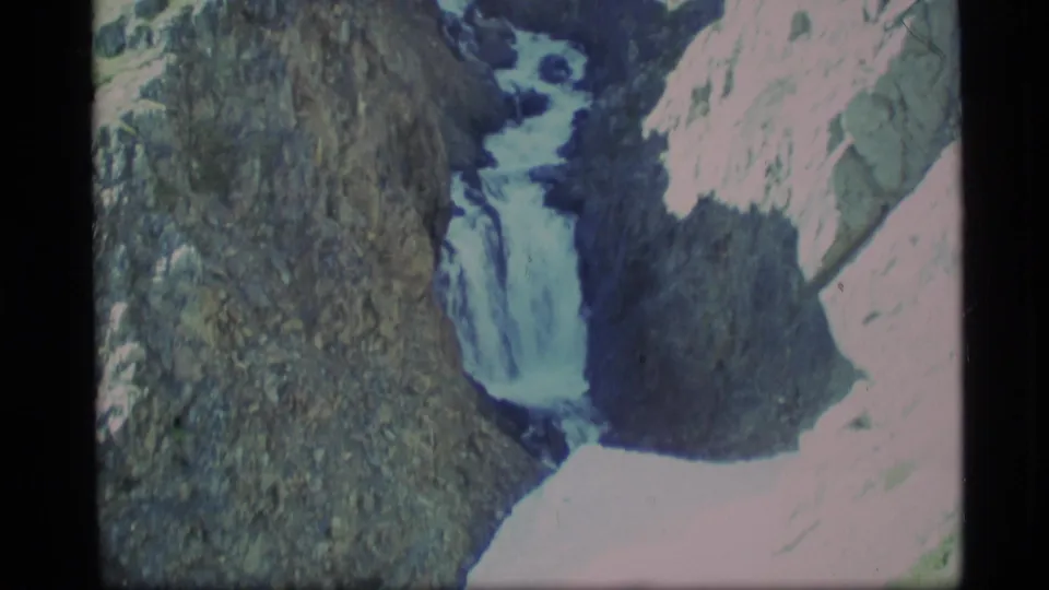 1977 Alaska 8mm view of a waterfall dropping past dark, textured rock with bright overexposed stone nearby.