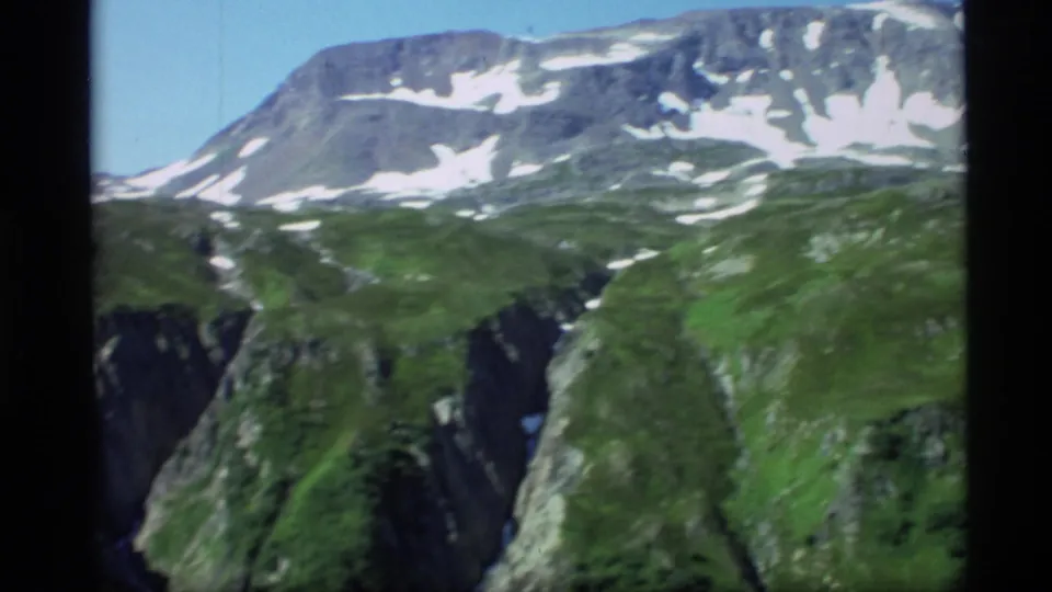 1977 Alaska 8mm frame showing a snow-dusted mountain ridge above green hills and a steep canyon.