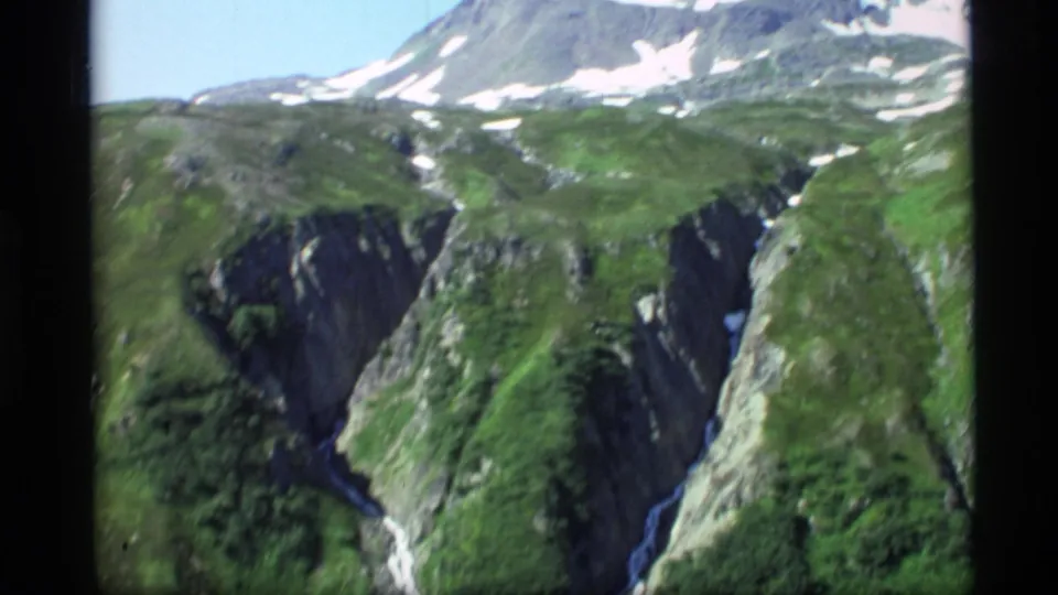 Vintage 8mm Alaska landscape view of green canyon walls below a snow-patched mountain ridge.
