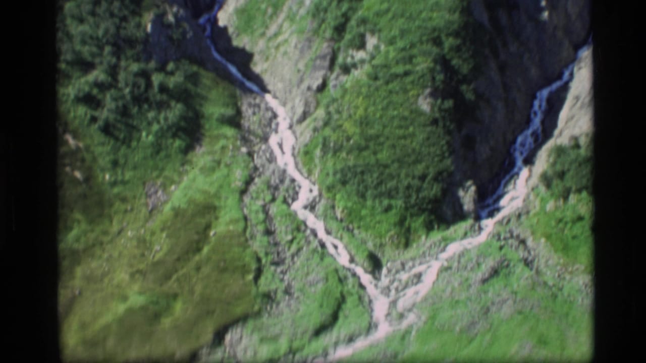 1977 Alaska 8mm frame with a high view over green alpine slopes, dark tree patches, and a pale winding ravine or trail.