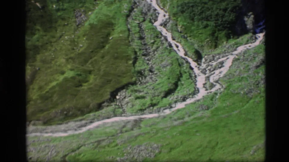 1977 Alaska 8mm frame showing a green valley floor, rocky ground, and a pale winding trail or streambed below steep slopes.
