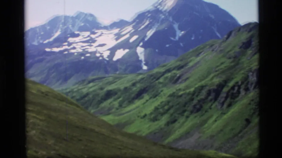 1977 Alaska 8mm frame showing snow-streaked mountain peaks beyond green ridges and a broad alpine valley.