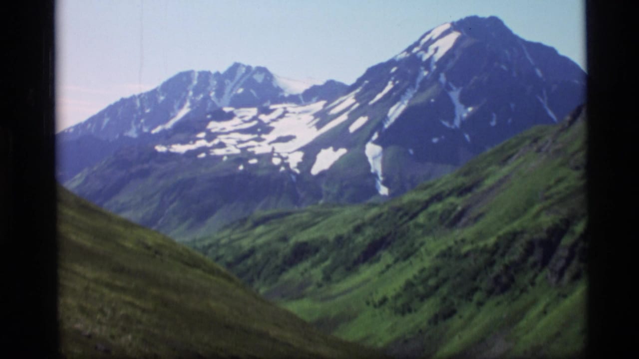 1977 Alaska 8mm frame showing snow-streaked mountain peaks rising above green alpine slopes under a pale sky.