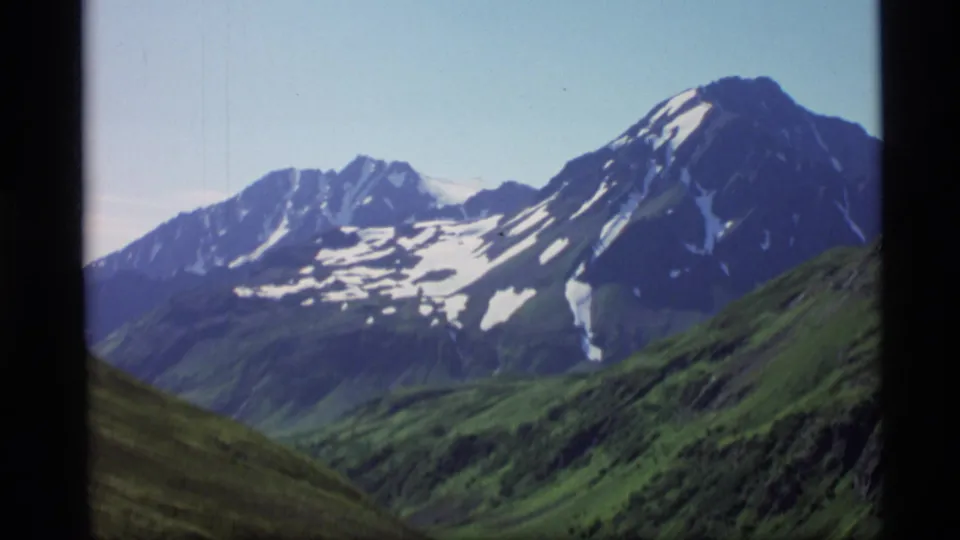 1977 Alaska home-movie frame of a wide mountain view with lingering snow on ridges and green slopes in the foreground.
