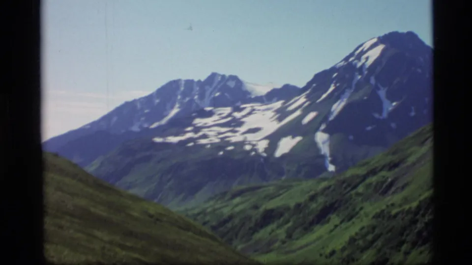 1977 Alaska 8mm frame looking toward snow-patched mountains beyond green valley slopes, with visible film borders.