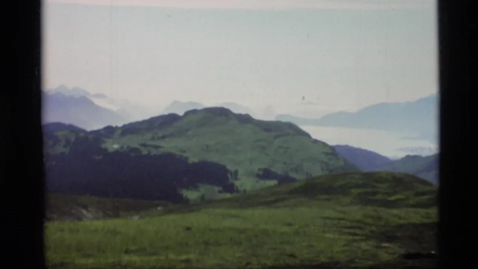 1977 Alaska 8mm frame showing green hills, dark forest patches, distant hazy mountains, and a possible body of water.
