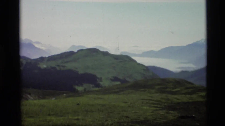 1977 Alaska 8mm frame showing rolling green hills, distant inlet water, mountain ridges, and dark window-frame edges.