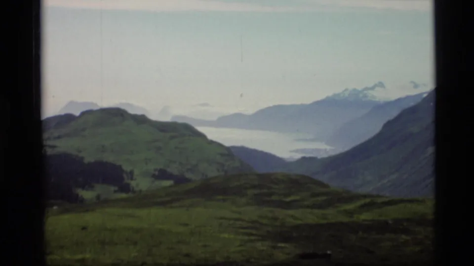 1977 Alaska 8mm view across green hills toward a pale inlet and layered mountain ridges under a light sky.