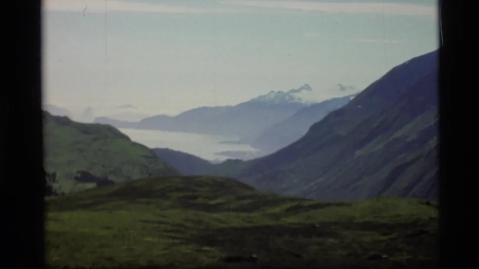 1977 Alaska 8mm frame with dark green hills, a distant inlet, and snow-capped mountain peaks partly softened by haze.
