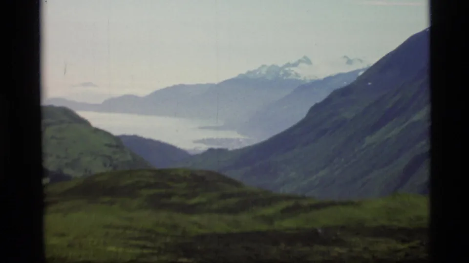 1977 Alaska 8mm frame showing a green hillside foreground, distant water, steep mountain slopes, and snow-topped peaks.