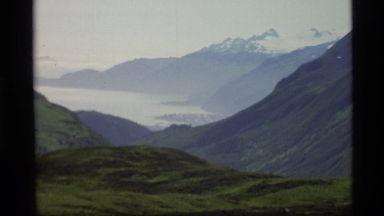 1977 Alaska 8mm frame showing green hills, a mountain valley, distant pale water, and snow-capped peaks in haze.