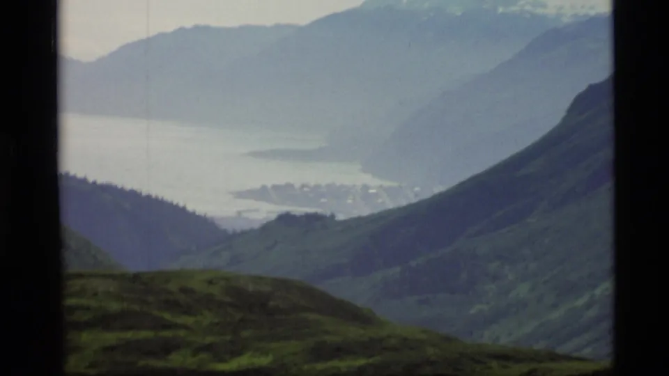 1977 Alaska home-movie frame with steep green slopes, hazy bay water, and a small coastal town far below.