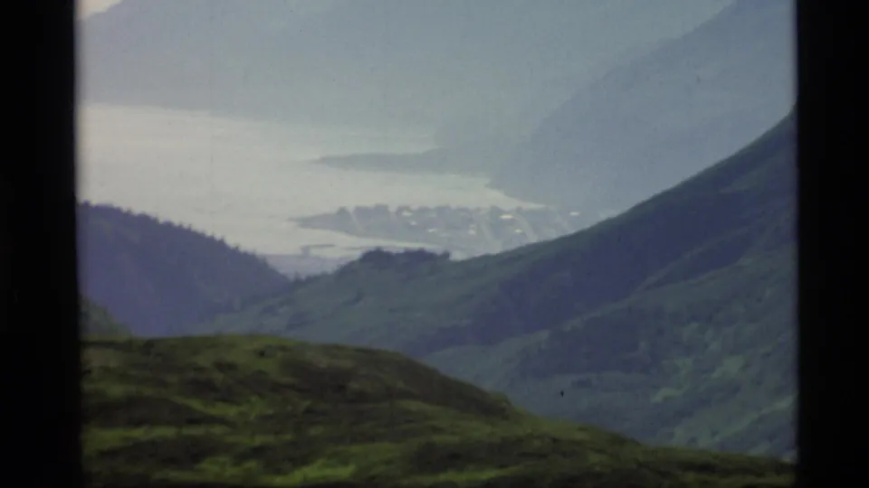 1977 Alaska 8mm view of a hazy bay and distant shoreline framed by dark green ridges and foreground hillside.