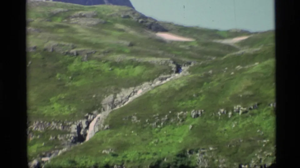 1977 Alaska 8mm frame showing a rocky green hillside with a narrow stream or gully cutting through the slope.