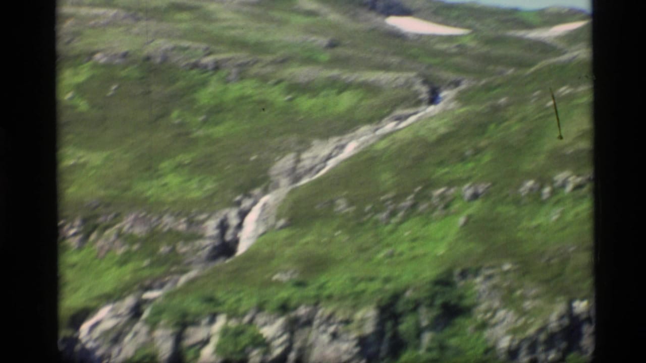 1977 Alaska 8mm frame showing a steep green hillside, exposed rock, and a narrow stream descending through a rugged ravine.